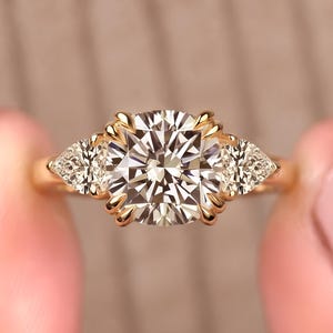 Pode incluir: Um anel de ouro com uma grande pedra central de corte brilhante redondo presa por garras, ladeada por duas pedras laterais menores em forma de pera. A banda é feita de ouro amarelo polido, criando um design clássico de três pedras.