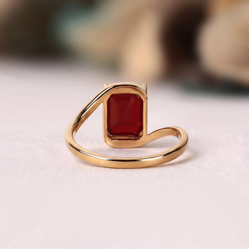 Rear angled view of a ruby red emerald cut gemstone ring highlighting the open back of the rectangular setting and sculptural yellow gold band.