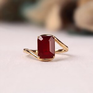 Front macro photograph of a deep ruby red emerald cut gemstone solitaire ring in polished yellow gold with graceful asymmetrical bypass shoulders and a four prong setting.