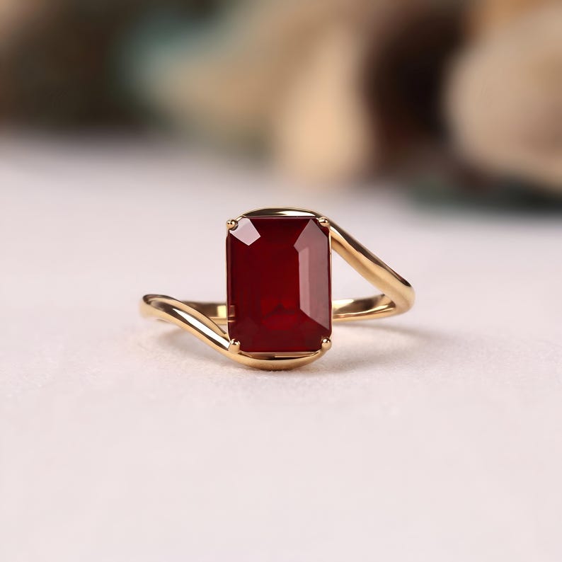 Front macro photograph of a deep ruby red emerald cut gemstone solitaire ring in polished yellow gold with graceful asymmetrical bypass shoulders and a four prong setting.