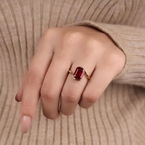 Lifestyle photograph of a woman wearing a ruby red rectangular gemstone ring with an asymmetrical yellow gold setting, styled against a soft beige sweater.