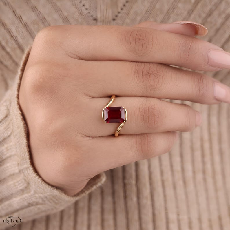 Angled hand modeling photograph of an emerald cut ruby red gemstone solitaire ring with a polished yellow gold bypass design and smooth curved shoulders.
