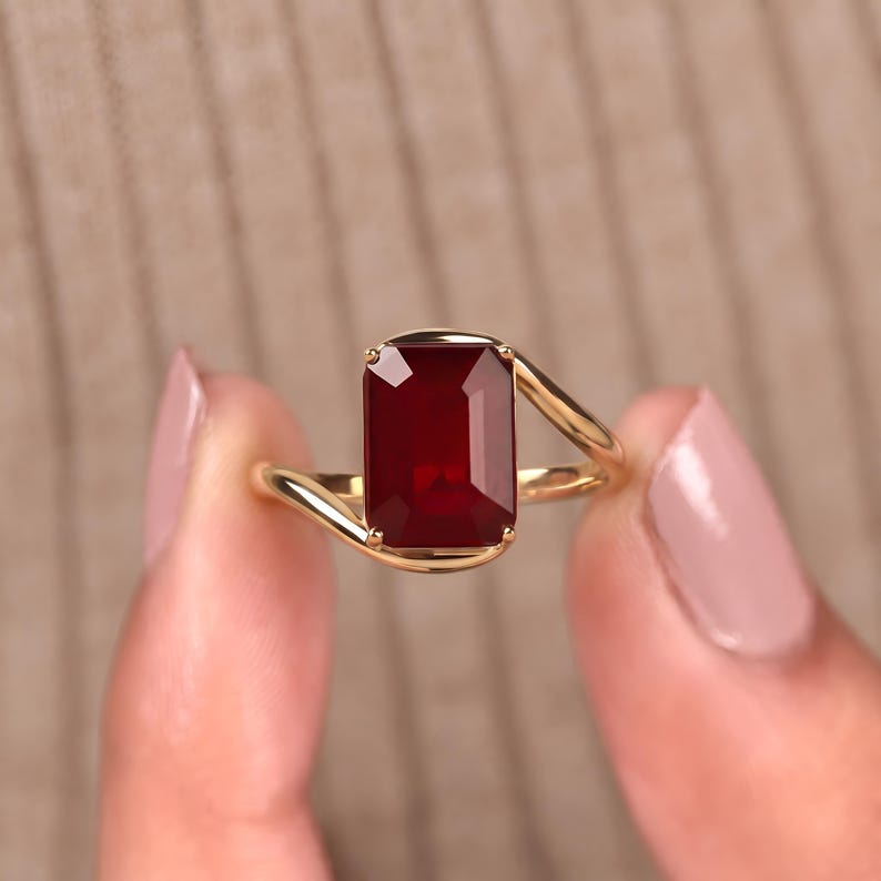 Close-up of a deep ruby red emerald cut gemstone ring worn on a woman's finger, featuring a yellow gold asymmetrical band and a bold rectangular center stone.