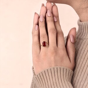 Macro photograph of a deep ruby red emerald cut gemstone ring held between two fingers, showing its clipped corners, four gold prongs and modern bypass band.