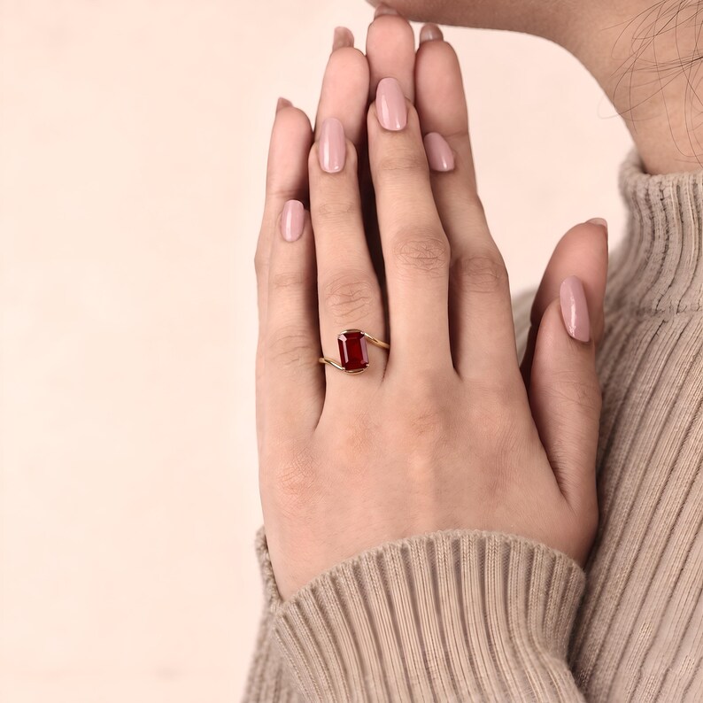 Macro photograph of a deep ruby red emerald cut gemstone ring held between two fingers, showing its clipped corners, four gold prongs and modern bypass band.