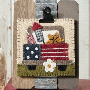 May include: A felt wall hanging featuring a red, white, and blue truck filled with fireworks. The truck has a white flower on the front and is sitting on a green field. The wall hanging is framed in a rustic wood frame.
