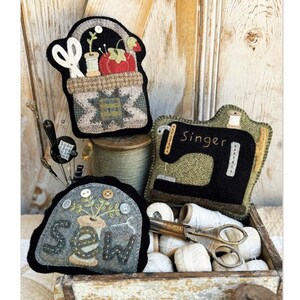 May include: Three felt ornaments in a rustic wooden box. The ornaments are a sewing basket, a sewing machine with the word "Singer" on it, and a spool of thread with the word "Sew" on it.