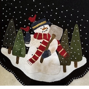 May include: A black felt banner with a winter scene. A snowman with a red scarf and hat holds a shovel. Green trees and red birds are in the snow. White dots represent falling snow. The banner is trimmed with white stitching.
