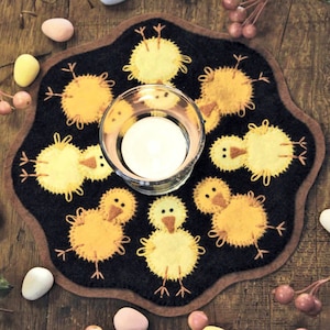 May include: A black felt coaster with a brown border features a circle of yellow chicks with brown eyes and beaks. The coaster is decorated with a clear glass votive holder in the center.