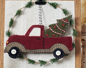 May include: A red truck with a green Christmas tree in the back, hanging from a wooden frame with a green wreath. The truck is made of felt and has a white button for a wheel. The background is a page of text.