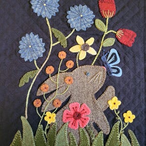 May include: A quilted textile featuring a whimsical design. A brown rabbit is surrounded by colorful felt flowers, including blue, red, yellow, and pink blooms. A blue butterfly rests near the rabbit. The background is a dark blue fabric.