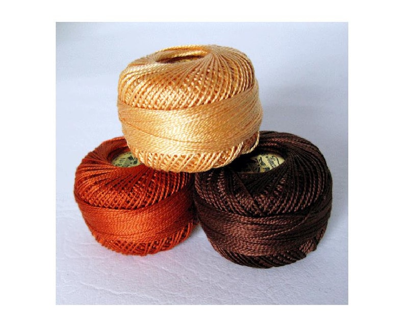 May include: Three spools of embroidery floss in orange, brown, and beige colors. The spools are stacked on top of each other.