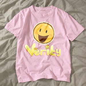 Puede incluir: Una camiseta de manga corta de color rosa claro con un gráfico de una cara sonriente circular amarilla con una boca ancha de color marrón oscuro. Debajo de la cara sonriente, la palabra Verity está impresa en una fuente amarilla estilizada con una pequeña estrella sobre la letra i.