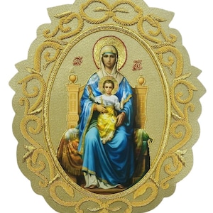 May include: An oval-shaped religious icon featuring a central image of a figure in a blue robe holding a child in a white and yellow garment, both seated on a throne. The image is surrounded by an ornate, raised gold-colored border with scrollwork.