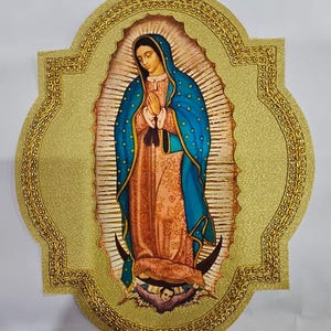 Może przedstawiać: Dekoracyjna tabliczka ścienna z wizerunkiem Matki Boskiej z Guadalupe. Centralna postać ma na sobie niebieski płaszcz w złote gwiazdy na różowej szacie, stoi na półksiężycu podtrzymywanym przez anioła, całość w złotej, brokatowej ramce.