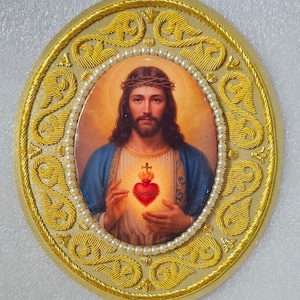 Puede incluir: Una placa de pared ovalada que presenta un retrato central de Jesucristo con una corona de espinas y un Sagrado Corazón brillante. La imagen está bordeada por un anillo de pequeñas perlas blancas y un marco exterior de bordado metálico dorado.