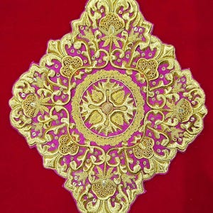 Puede incluir: Un parche bordado en forma de diamante con un intrincado trabajo de hilo metálico dorado sobre una base de tela magenta vibrante. El diseño incluye un motivo circular central rodeado de elaborados patrones florales y lentejuelas.