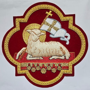 Può includere: Una toppa ricamata raffigurante un agnello color crema che riposa su una superficie rossa. L'agnello tiene una bandiera bianca con una croce dorata. Un'aureola dorata si trova dietro la testa. La toppa ha uno sfondo rosso scuro e un bordo dorato.