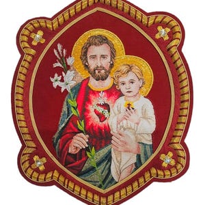 Puede incluir: Un tapiz decorativo bordado que representa a San José sosteniendo al niño Jesús. Las figuras se sitúan sobre un fondo rojo intenso con halos dorados, rodeadas por un borde dorado adornado con cuatro cristales decorativos.