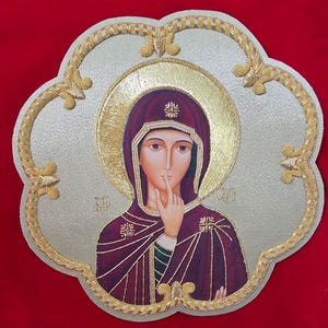 May include: A decorative wall plaque featuring a central religious icon of a figure in a deep purple hooded garment with a gold halo, set against a textured silver background with a scalloped edge and gold embroidered trim detailing.