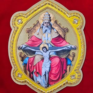Op de afbeelding: Een religieuze icoon met een centrale afbeelding van een bebaarde figuur met een pauselijke tiara, zittend op een troon en een kruis vasthoudend met een gekruisigde figuur. Het beeld is omlijst door een goudkleurige rand met decoratieve koordafwerking.