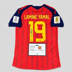 May include: A red football shirt with dark blue sleeves, featuring the name LAMINE YAMAL and the number 19 in yellow. The shirt includes a black signature on the number, a United For Peace patch, a 2026 World Cup patch, and a certificate of authenticity.