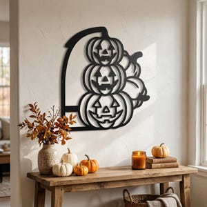 May include: A black metal wall art piece featuring a vertical stack of three carved jack-o'-lanterns with triangular eyes and jagged mouths, framed by a curved arch design, mounted on a light-colored wall above a wooden console table.
