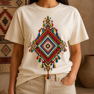 Peut inclure: Un t-shirt à manches courtes de couleur crème arborant un grand motif central en forme de losange. Le design se compose de motifs géométriques complexes en rouge, bleu, vert et jaune, agrémentés de petites franges suspendues.