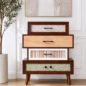 May include: A four-drawer chest with a dark wood frame and tapered legs. The top drawer features a chevron pattern, the second is plain light wood, the third has vertical stripes, and the bottom drawer displays a light green panel with gold scrollwork.