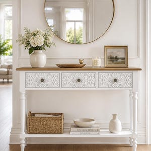 May include: A white console table with a natural wood top and three drawers featuring intricate floral carvings and small round knobs. The table has four turned legs and a lower shelf holding a woven basket, a ceramic bowl, two books, and a vase.