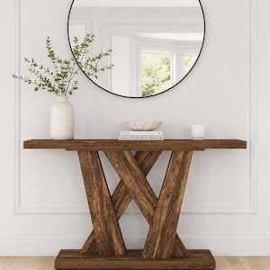 May include: A dark brown wooden console table with a rectangular top and a base featuring two intersecting diagonal supports. The table is topped with a white ceramic vase containing green foliage, a shallow bowl, two stacked books, and a white candle.