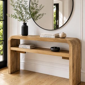 May include: A light oak console table with a curved waterfall design, featuring a lower shelf. The top surface holds a black ribbed vase with greenery, two stacked books titled Kinfolk Home and Live Beautifully, and a small beige ceramic bowl.