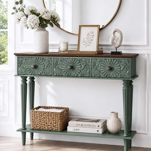 May include: A sage green console table with a dark wood top, featuring three drawers with carved floral circular patterns and round knobs. The table has four fluted legs and a lower shelf holding a woven basket, two books, and a small ceramic vase.