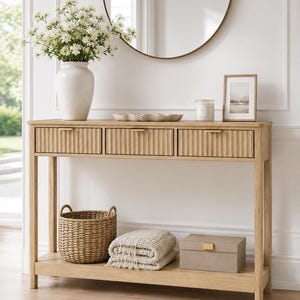 May include: A light wood console table featuring three drawers with vertical fluted fronts and thin gold-toned metal handles. The table has a lower shelf holding a woven basket, a folded beige textile, and a rectangular box with a gold latch.