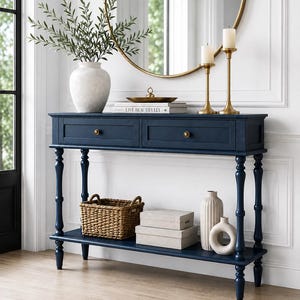 May include: A navy blue console table with two drawers, brass knobs, and four turned legs. The table features a lower shelf holding a woven basket, two stacked rectangular boxes, a tall ribbed vase, and a circular ceramic sculpture.
