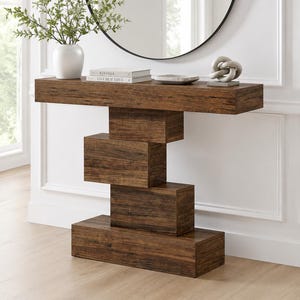 May include: A dark brown wood-grain console table featuring a rectangular top and a base constructed from four stacked, offset rectangular blocks. The table surface holds a white vase with greenery, two stacked books, a small bowl, and a knot sculpture.