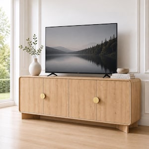 May include: A light wood media console with rounded corners and four cabinet doors featuring circular gold hardware. The console supports a flat-screen television displaying a landscape image of a lake and forest, with a white vase and books nearby.