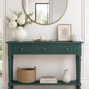 May include: A dark teal console table featuring three drawers with intricate carved floral patterns and round knobs. The table has four turned legs and a lower shelf holding a woven basket, two stacked books, and a small white ceramic vase.