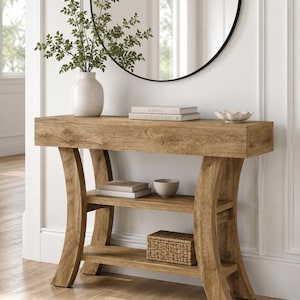May include: A light brown wooden console table with a rectangular top, two lower shelves, and curved legs. The table holds a white vase with green foliage, two stacked books, a small white bowl, a small gray bowl, and a woven basket.