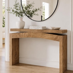 May include: A light brown wooden console table with a thick rectangular top resting on an arched base. On the table sits a white vase with green foliage, two stacked books titled Live Beautifully and Calderone, and a shallow white bowl.