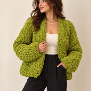 Puede incluir: Un cárdigan abierto de color verde lima, tejido con hilo grueso y suave que presenta una textura de bucles pronunciada. La prenda cuenta con mangas largas y anchas, con un corte holgado, combinada con una camiseta blanca y pantalones negros.