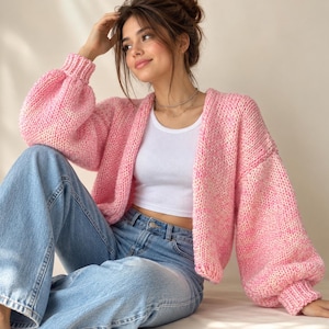May include: A chunky knit cardigan in a variegated pink and white yarn featuring long, voluminous balloon sleeves with ribbed cuffs, an open front design, and a relaxed fit, worn over a simple white ribbed tank top and light blue denim jeans.