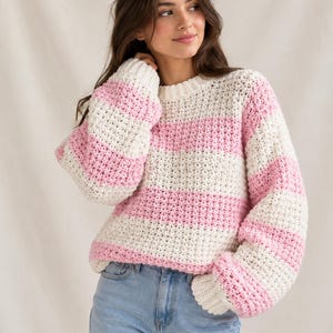 May include: A chunky knit sweater featuring horizontal stripes in cream and light pink. The garment has a round neckline, long sleeves with ribbed cuffs, and a textured, thick yarn construction that provides a voluminous, relaxed fit throughout.