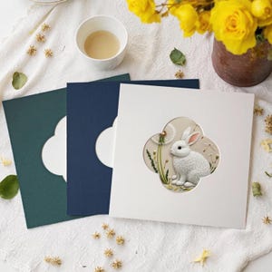 Puede incluir: Tres tarjetas de felicitación cuadradas con ventanas troqueladas en forma de trébol. La tarjeta superior es blanca y presenta una ilustración detallada de un conejo blanco sentado junto a un diente de león. La tarjeta central es azul oscuro y la inferior es verde oscuro.
