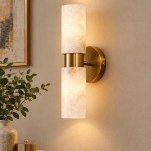 May include: A wall-mounted light fixture featuring two cylindrical, translucent, cream-colored stone shades with marbled patterns. The shades are separated and connected by a central brushed brass metal band and attached to a circular brass wall plate.