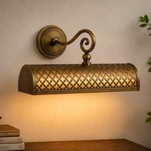 May include: A wall-mounted picture light in an antique brass finish featuring a cylindrical shade with a repeating quatrefoil cutout pattern. The fixture has a curved arm extending from a circular base and emits a warm downward glow.