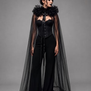 May include: A black floor-length sheer tulle cape featuring a high ruffled collar with a satin ribbon tie closure, worn over a black strapless corset top with front hook-and-eye fasteners and matching black high-waisted wide-leg trousers.