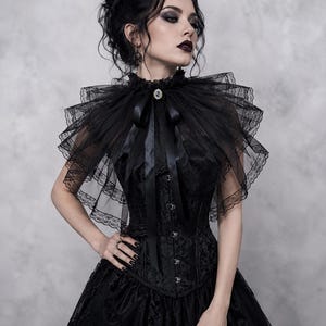 Könnte beinhalten: Ein schwarzes Korsettkleid mit einem mehrlagigen Tüll- und Spitzenkragen in Cape-Optik. Der Kragen wird mit einer schwarzen Satinbandschleife und einer ovalen Brosche in der Mitte gehalten. Das Oberteil besteht aus gemustertem Stoff.