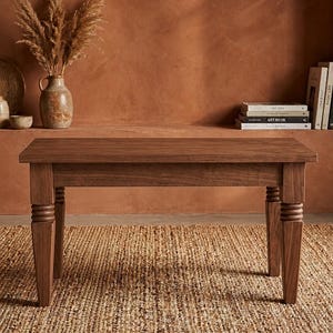 May include: A rectangular wooden bench featuring a smooth, flat top surface and four sturdy legs. Each leg is designed with a distinct turned, ribbed detail near the top, transitioning into a tapered shape that extends down to the floor.