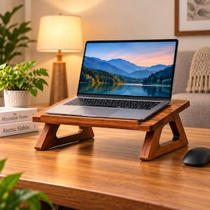 Może przedstawiać: Ciemnobrązowy drewniany stojak na laptopa z nachyloną powierzchnią i dwiema solidnymi bocznymi podporami, podtrzymujący srebrny laptop z obrazem gór i jeziora na ekranie, umieszczony na pasującej drewnianej powierzchni biurka.
